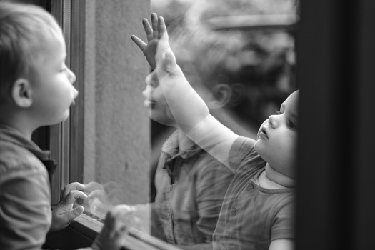 Young child with hand raised against window glass, reaching toward their reflection in black and white.