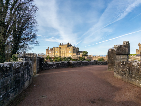 Stone fortification walls frame a golden-lit castle on a distant hillside under blue sky.