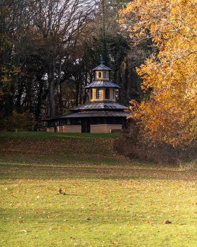 Octagonal gazebo with lit interior among autumn trees and fallen leaves on lawn.