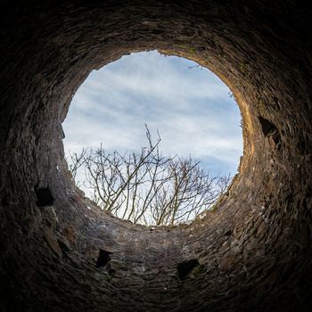 View through a circular stone tunnel opening onto bare branches and cloudy sky.