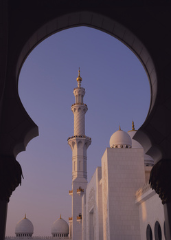 White mosque with tall minaret framed through dark stone archway at dusk.