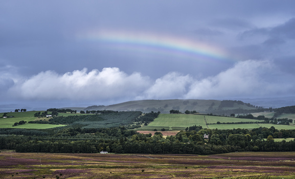 Rolling rural landscape with green fields, forested areas, and distant hills under dramatic cloudy sky with a faint rainbow.