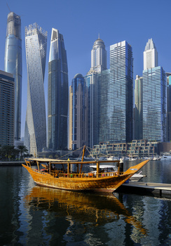 Traditional wooden dhow boat with golden-yellow sails on calm water, framed against towering modern skyscrapers under clear blue sky.