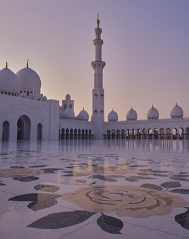 Grand mosque courtyard at dusk with ornate geometric floor patterns and illuminated minarets.