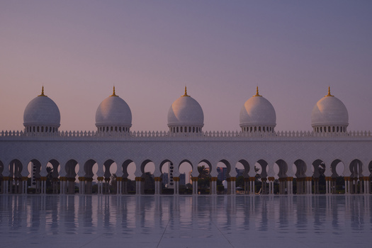 Five domes of a mosque silhouetted against a purple twilight sky, reflected in wet ground below.