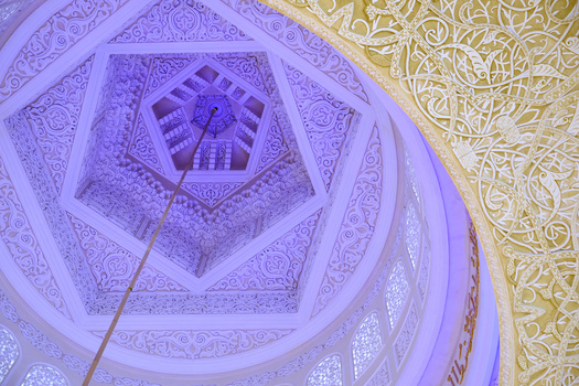 Upward view of ornate geometric ceiling with concentric hexagonal patterns and intricate carved details in purple and gold lighting.