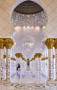 Symmetrical corridor with white marble columns, ornate gold capitals, and geometric floor patterns leading to a vanishing point.