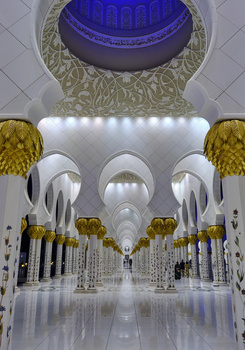 Interior corridor of a grand mosque with symmetrical white arches and golden decorative capitals.