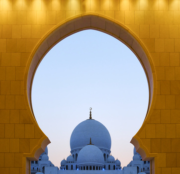 Blue-domed mosque framed through a large horseshoe arch in golden tile wall.