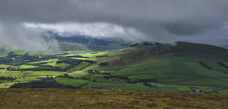 Rolling green valleys and hillsides under dramatic cloudy skies, with distant mountains shrouded in mist.