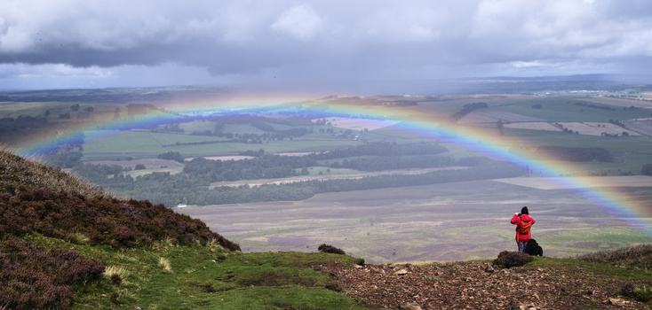 Photographer in red jacket capturing a dramatic double rainbow arcing over agricultural valley landscape.