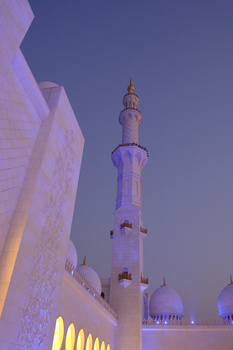 Illuminated minaret tower rising against twilight sky beside modern white architectural structure.