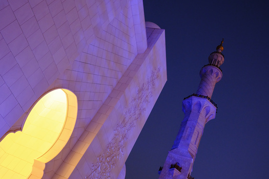 Nighttime architectural shot of illuminated modern building with glowing yellow circular element and minaret tower in blue light.