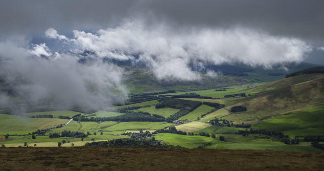 Dramatic cloudy sky over rolling green farmland valleys with scattered buildings and field divisions.