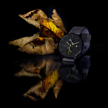 Black chronograph watch positioned next to dried autumn leaves on reflective black surface.