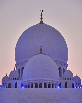 Illuminated white mosque with large domes and crescent finials at dusk.
