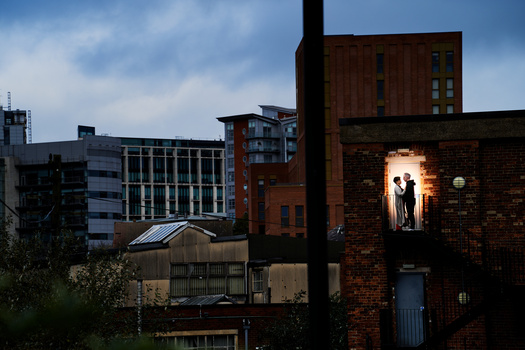 Silhouetted figures standing in a lit brick building doorway at dusk, surrounded by modern urban architecture.