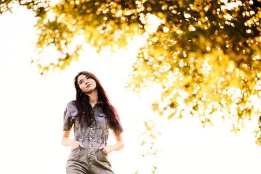 Woman in denim shirt looking upward beneath golden foliage backlit by bright sky.