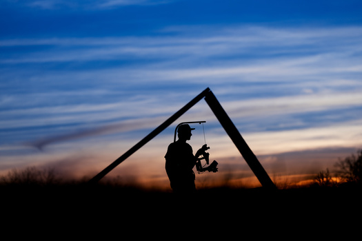 Silhouetted figure holding a lantern beneath an A-frame structure at sunset.