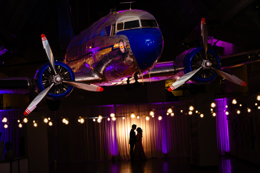 Silhouetted couple posing beneath a vintage aircraft suspended in dramatic blue and purple lighting.