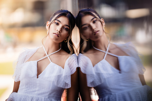 Two women in white off-shoulder garments pose back-to-back with heads tilted toward each other in a mirrored composition.