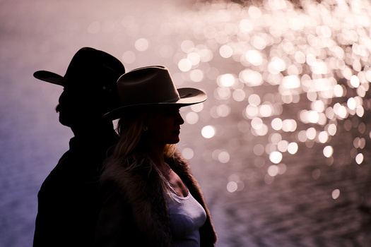 Two silhouetted figures wearing cowboy hats, backlit by bokeh lights reflecting off water.