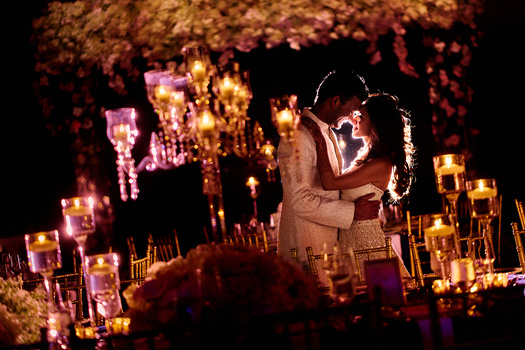 Bride and groom embrace under an ornate floral arch illuminated by warm golden candles at night.