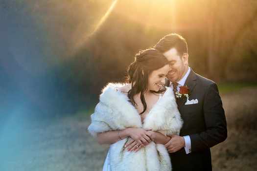 Bride and groom embracing outdoors during golden hour with warm sunlight streaming through trees.