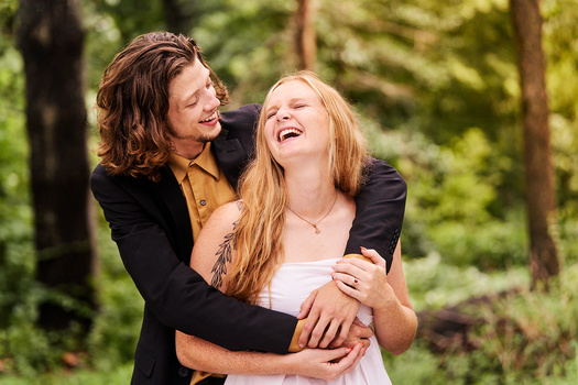 Two women embracing outdoors with trees in the background, both smiling and looking upward.