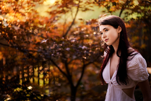Woman with long dark hair in white dress looking over shoulder with autumn foliage background.