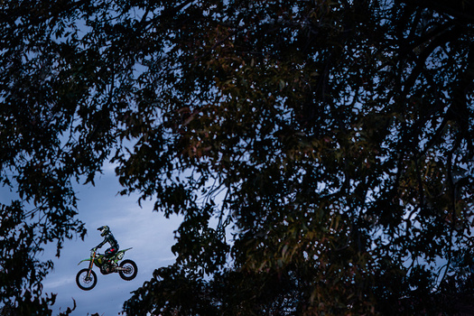 Motocross rider airborne on a dirt bike against a twilight sky framed by tree canopy.
