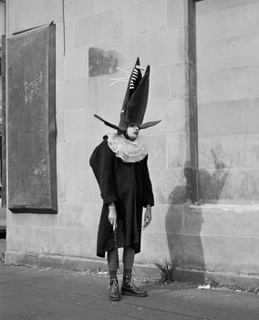 Person wearing dark coat with oversized black bird head obscuring their face, standing against white concrete wall.