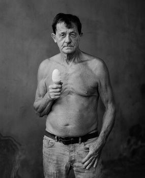 Shirtless middle-aged man with fist raised to chest against neutral gray background.