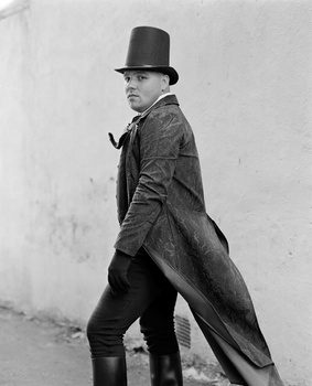 Man in top hat and overcoat standing in profile against plain wall.