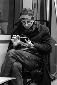 Man in cap and glasses sits on public transit, focused on handheld device in black and white.