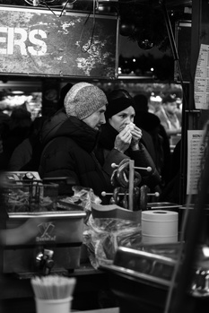 Man in winter hat and dark coat eating from a container on a city street at night.