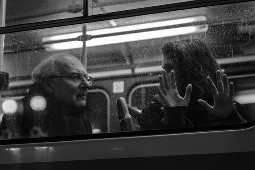 Two people visible through a bus window at night, one wearing glasses on the left, hands pressed against the glass.