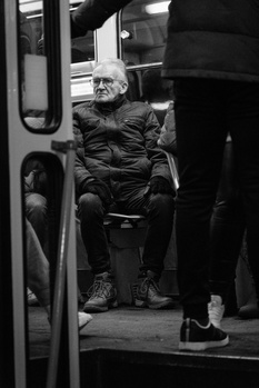 Elderly man seated inside a bus, wearing glasses and dark winter clothing, captured in black and white.