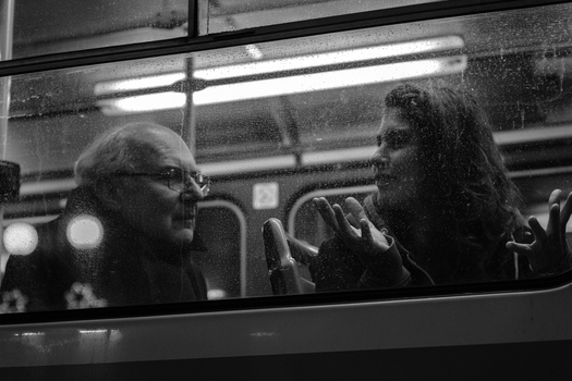 Two people conversing inside a vehicle, captured in black and white through the window.