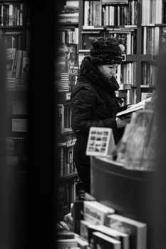 Person browsing books on library shelf, photographed through foreground obstruction in black and white.