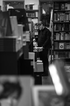 Person standing in a library or study room surrounded by bookshelves, photographed in black and white.