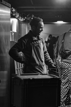 Man in jacket standing at table in dimly lit indoor venue, captured in black and white.