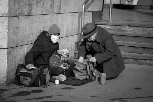 Two people sitting on pavement against a concrete wall, wearing winter coats and hats, sharing a meal from containers.