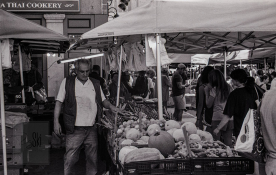 Vendor at outdoor market stall displaying fresh produce under tent canopy.