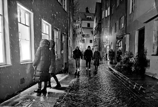 Wet cobblestone street at night with pedestrians walking past illuminated building facades.