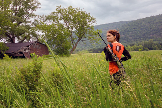 Woman in orange vest holding scythe in tall grass field with mountains and trees in background.