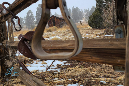 Rusted metal loop and chain in sharp focus with weathered ranch structures blurred in background.