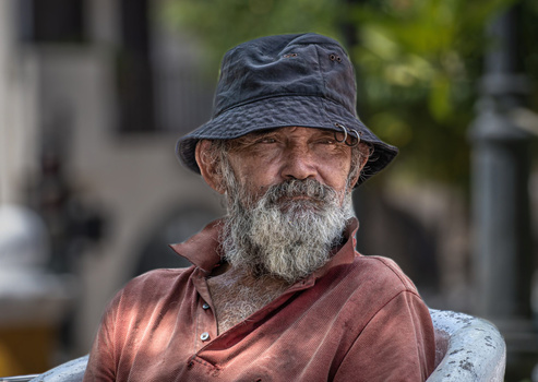 Older man with full gray beard wearing a rust-colored shirt and dark bucket hat, photographed outdoors with a soft-focused background.