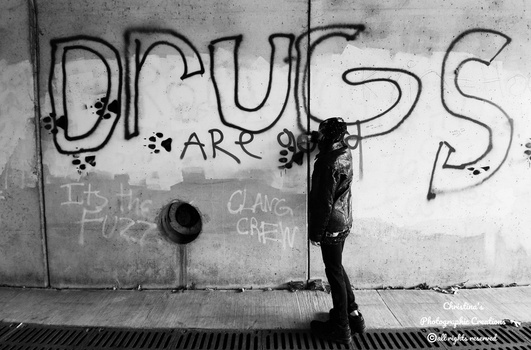 Person standing against concrete wall with large "DRUGS" graffiti text in black and white photograph.