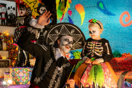 Adults and child in skeleton and skull face paint wearing black clothing posed against colorful Day of the Dead backdrop.